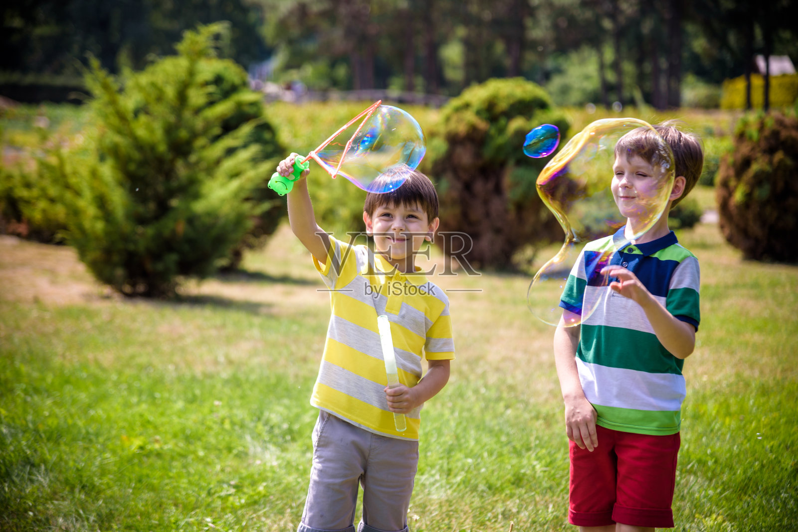男孩吹肥皂泡，而一个兴奋的孩子享受泡沫。快乐的少年和他的兄弟在公园里享受肥皂泡。童年快乐友谊理念照片摄影图片