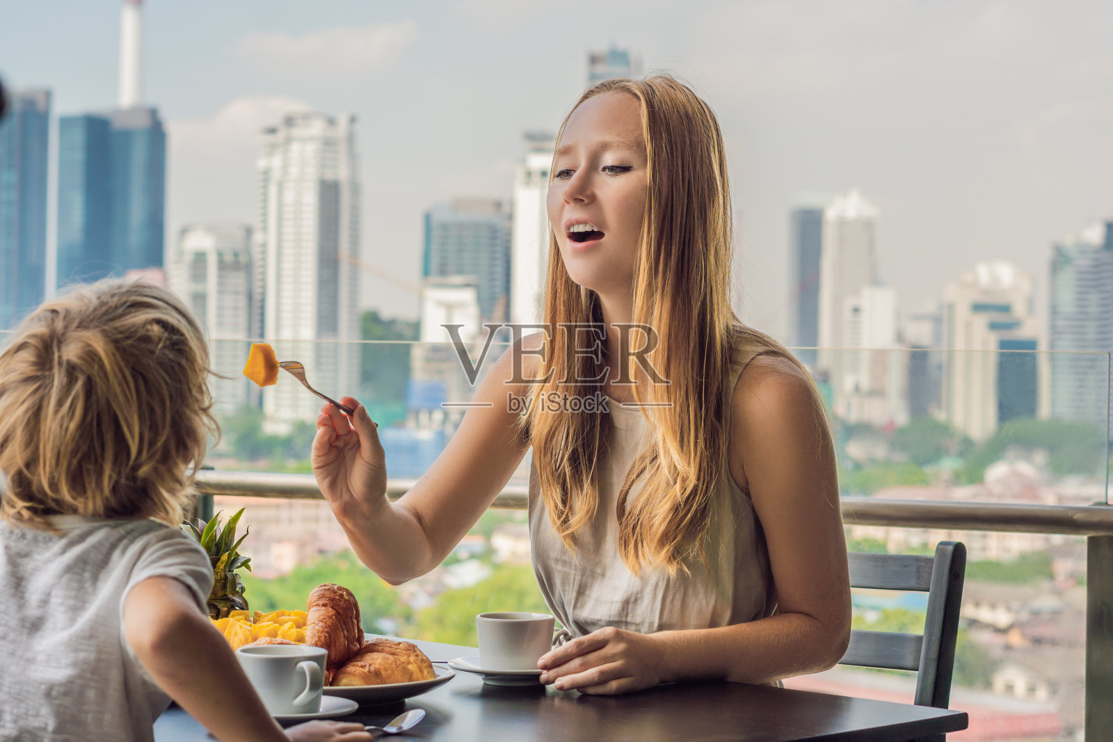 幸福的一家人在阳台上吃早餐。阳台上的早餐桌上放着咖啡、水果和牛角面包，背景是大城市照片摄影图片