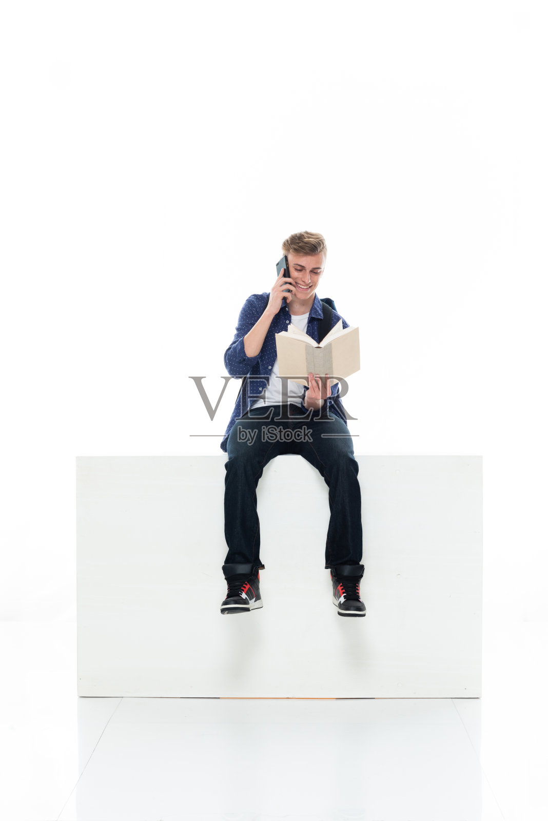 男大学生穿着牛仔裤在白色背景前休息，拿着书和使用手机照片摄影图片