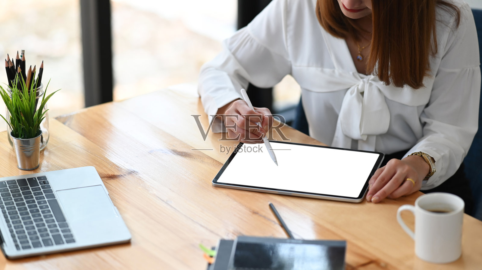 裁剪图像腰部以上的美丽女子作为秘书绘制一个商业计划在白色空白屏幕笔记本电脑，而坐在现代木制桌子与有序的办公室为背景。照片摄影图片