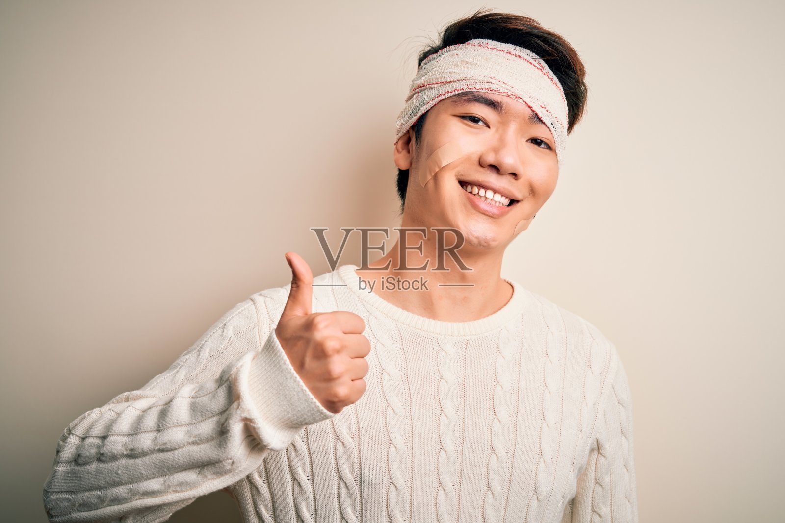 一名年轻帅气的中国男子因意外受伤，头上缠着绷带，用手做快乐的竖起大拇指的手势。赞许的表情看着镜头，表示成功。照片摄影图片