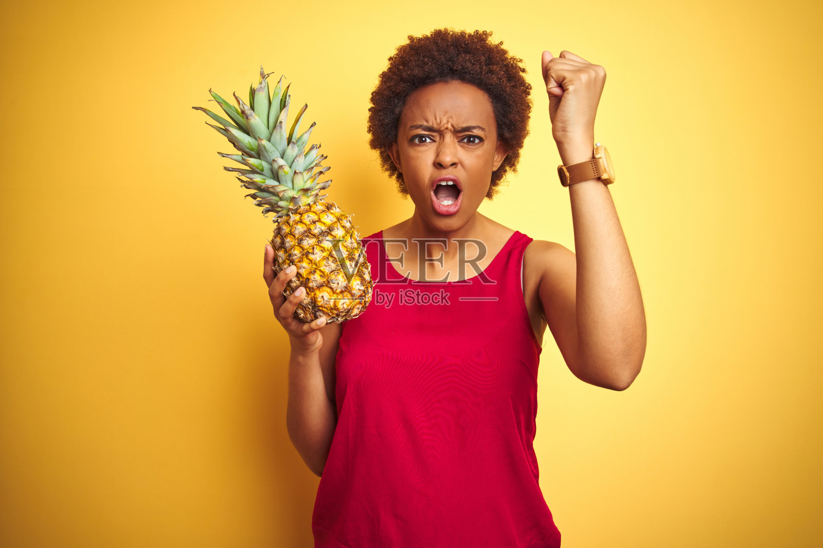 非洲裔美国妇女拿着热带菠萝在黄色孤立的背景生气和沮丧的大喊与愤怒，疯狂和大喊与高举的手，愤怒的概念照片摄影图片