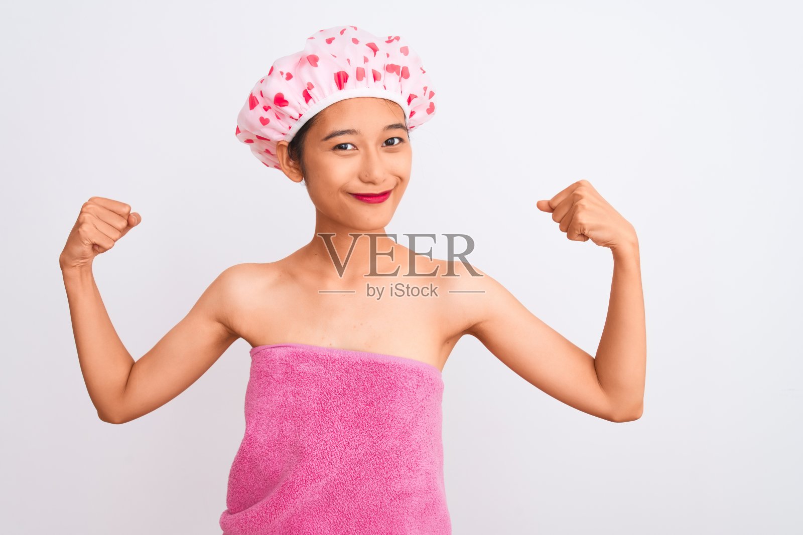 年轻的中国妇女穿着浴巾和帽子浴在孤立的白色背景显示手臂肌肉自豪地微笑。健康的概念。照片摄影图片