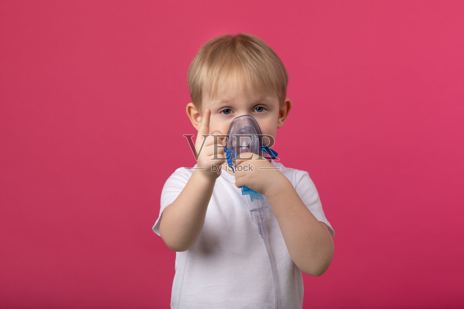 一个金发碧眼的孩子，嘴里衔着一个吸入器，用手指指着一个纯粉色的背景。照片摄影图片