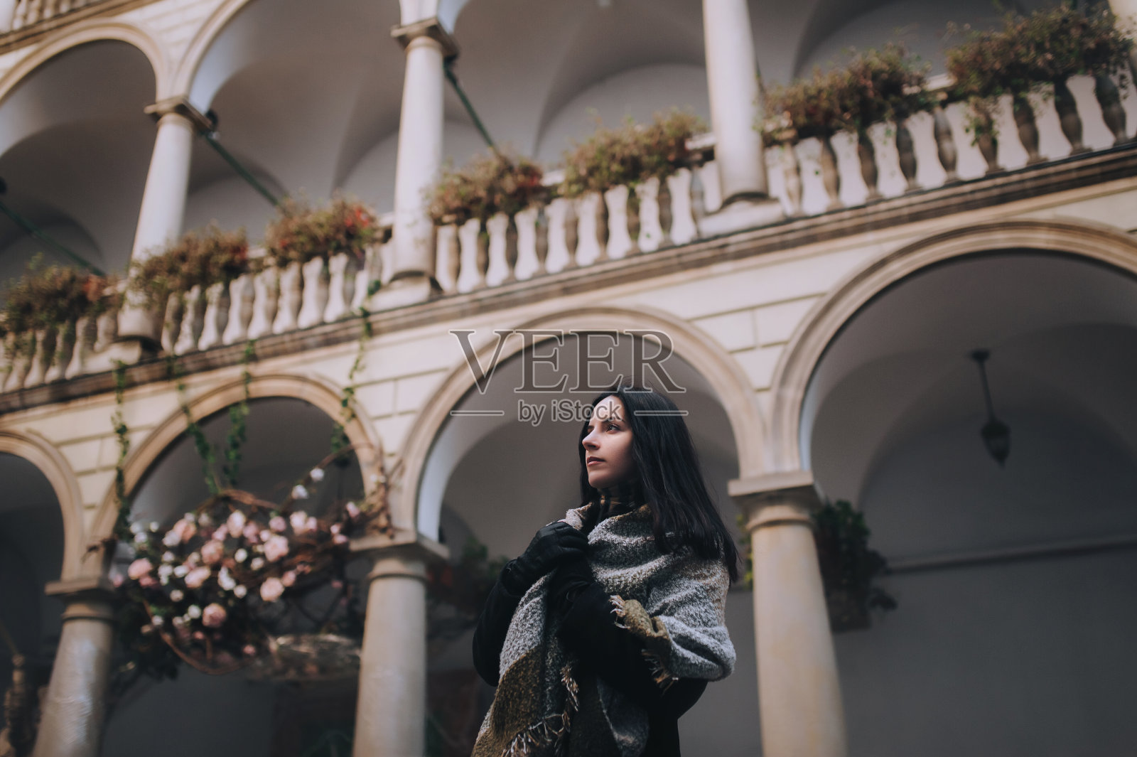 一个年轻的白种人黑发女孩，穿着深色的秋日外套，裹着宽大的围巾，站在灰色的柱廊背景上，看起来很直。利沃夫，意大利庭院，乌克兰。照片摄影图片