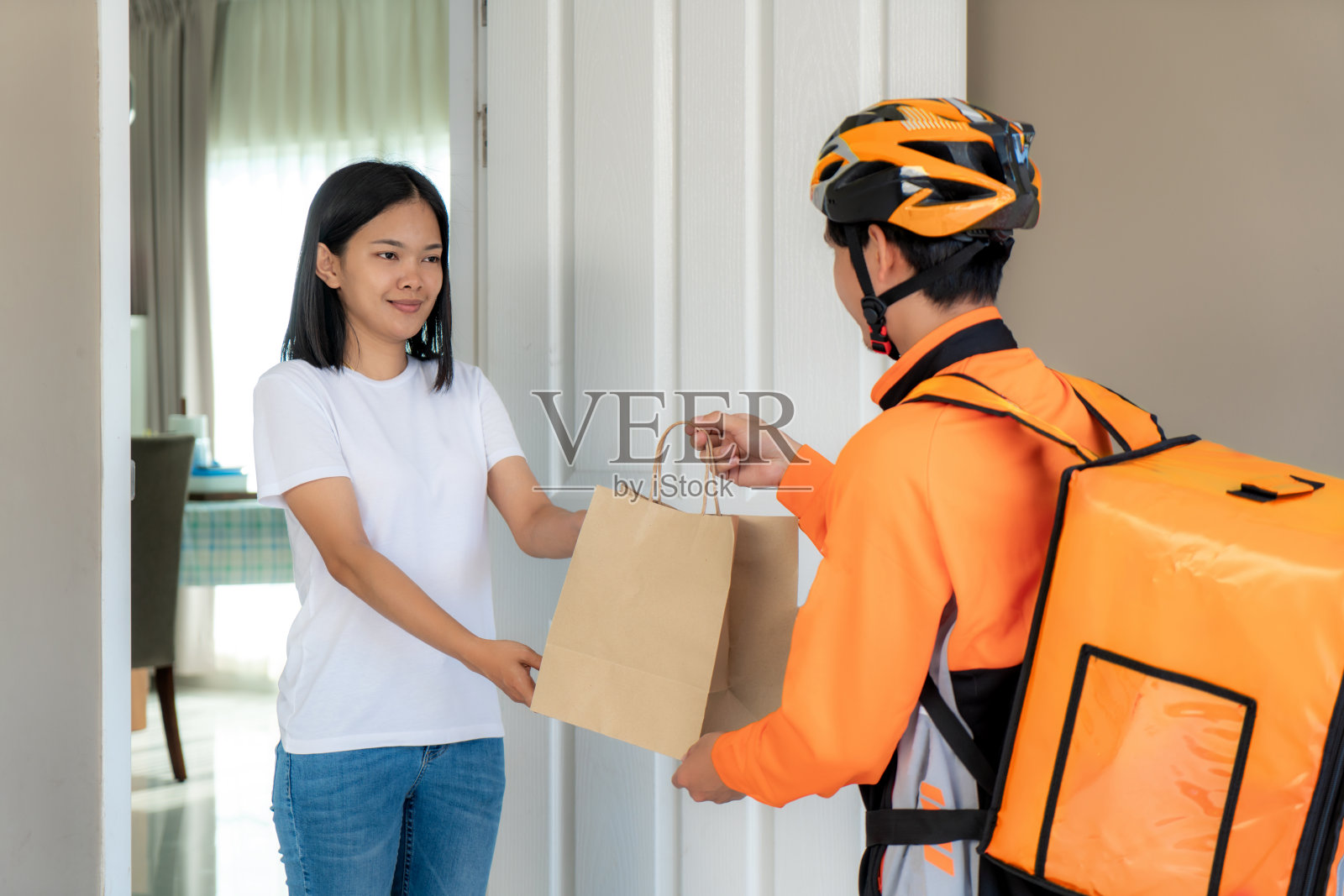 亚洲男子快递在自行车送食物在橙色制服微笑和食品袋在前面的房子和亚洲妇女接受快递员送的盒子。照片摄影图片