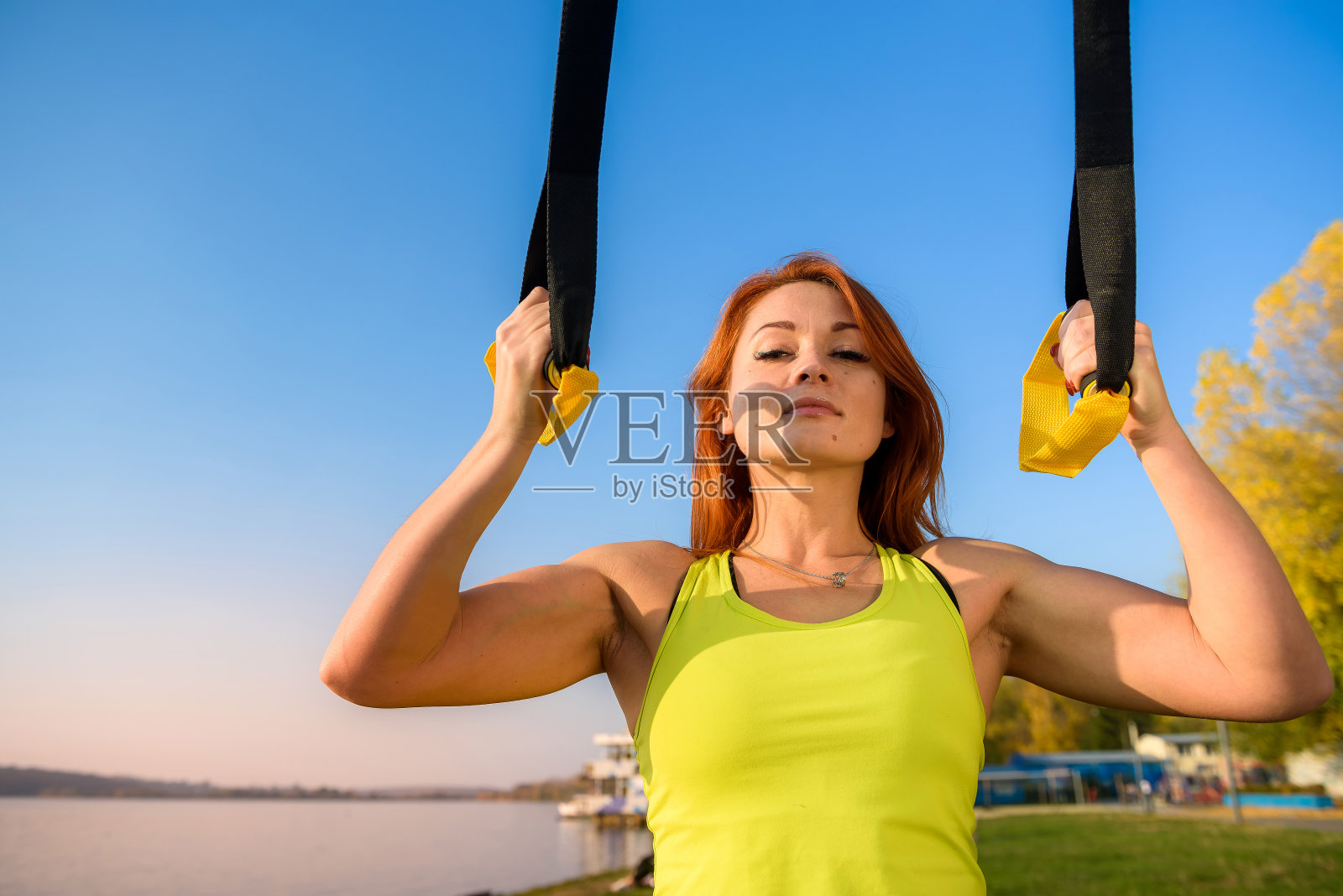 年轻苗条的女人做悬浮训练与健身带在户外附近的湖在白天。健康的生活方式。照片摄影图片