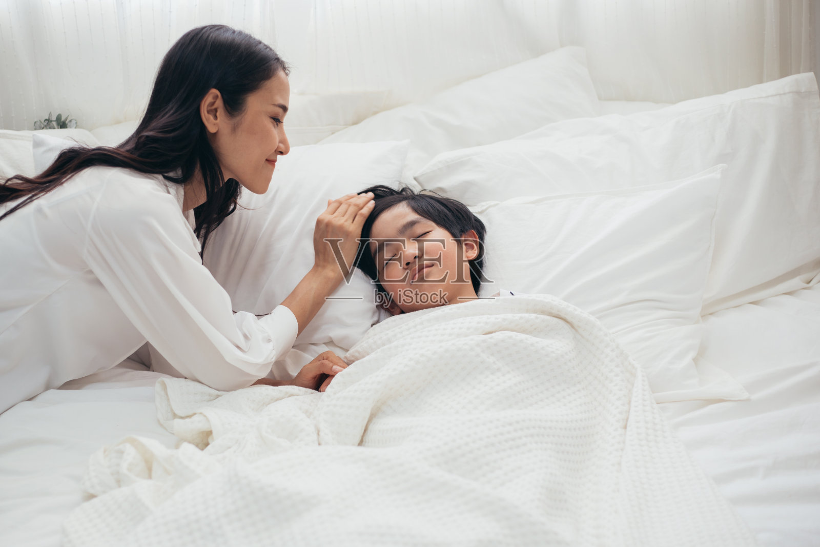 年轻的亚洲母亲用爱和微笑照顾看着她的儿子睡觉，生病的男孩睡在床上，妇女担心冠状流感，医疗保健和预防流行性感冒病毒的概念照片摄影图片