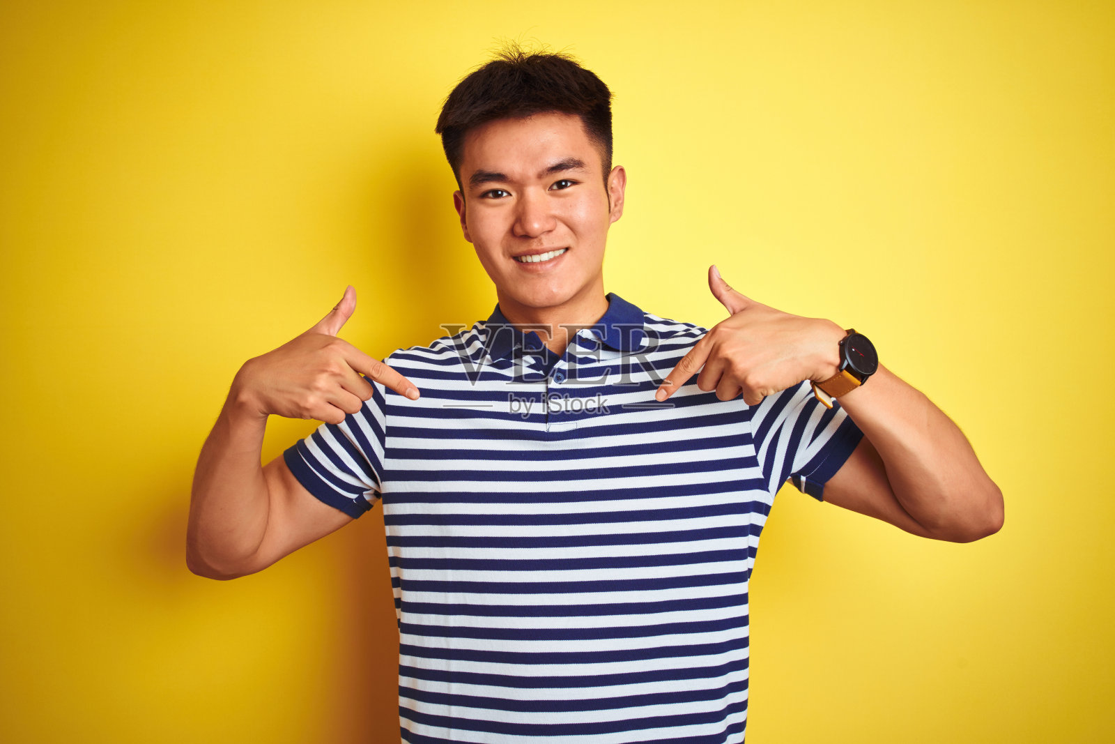 年轻的亚洲华人男子穿着条纹马球站在孤立的黄色背景看起来自信，脸上带着微笑，用手指指着自己骄傲和幸福。照片摄影图片