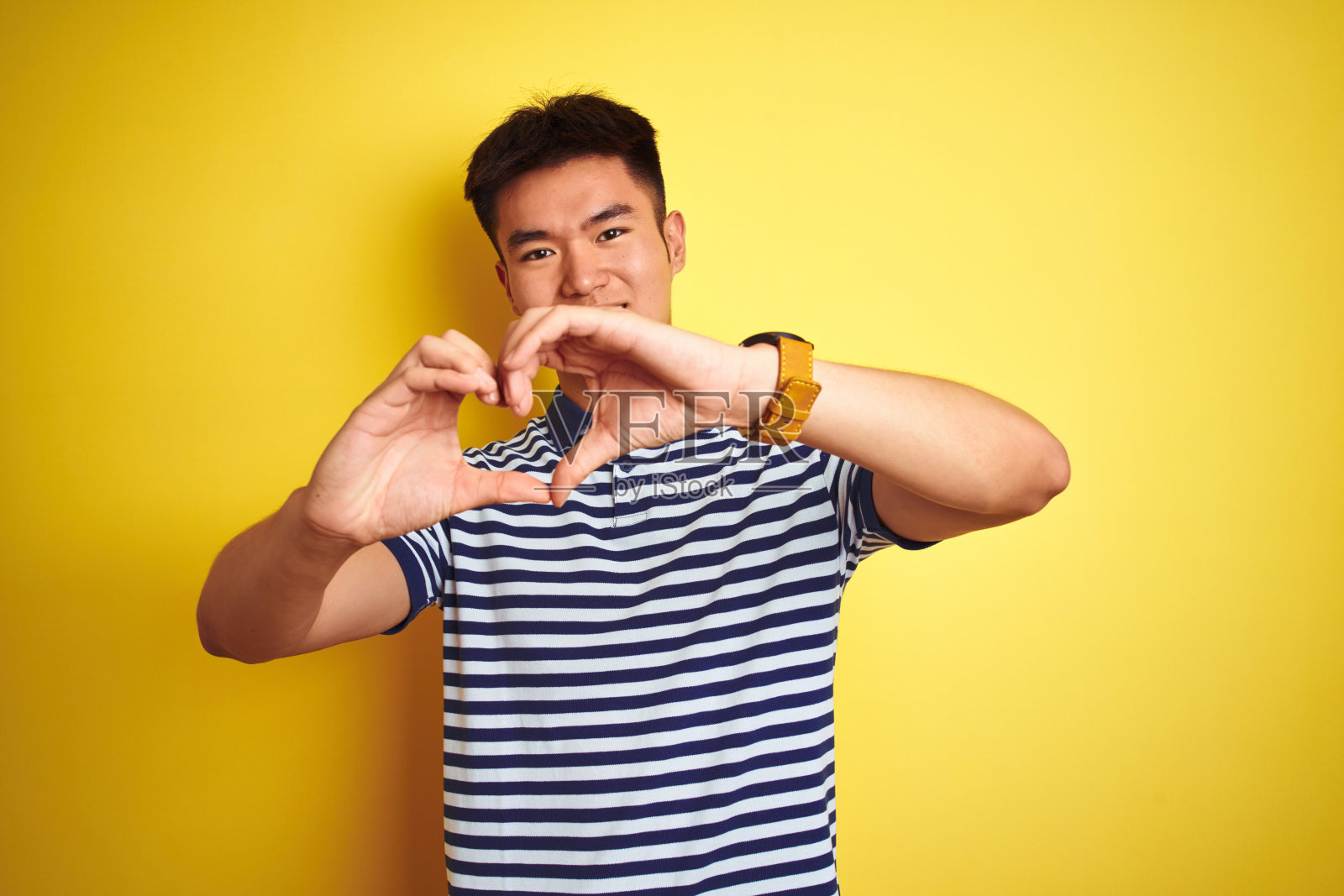 年轻的亚洲华人男子穿着条纹马球站在孤立的黄色背景在爱做心形符号形状的手微笑。浪漫的概念。照片摄影图片