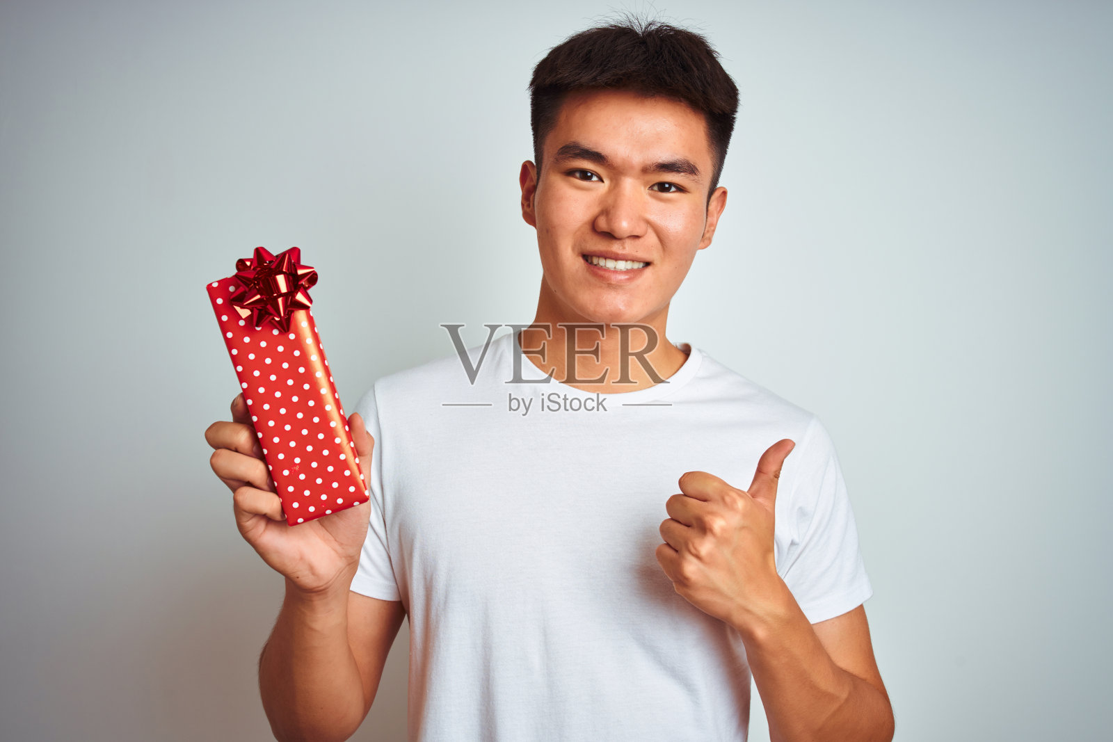 年轻的亚洲华人男子捧着情人节礼物站在孤立的白色背景下，开心地笑着做ok手势，用手指竖起大拇指，优秀的手势照片摄影图片