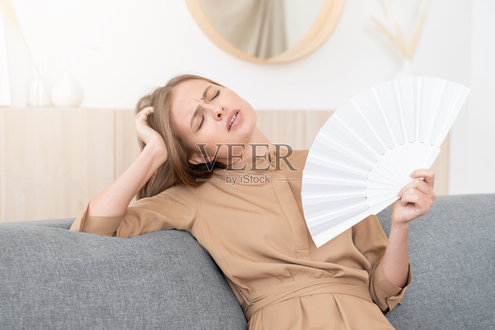 女孩坐在家里的灰色沙发上，经历着炎热的夏天，扇着风来应对炎热，看起来很烦恼和烦恼照片摄影图片