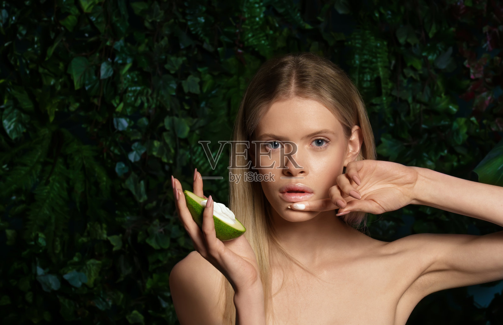 美丽的裸体模特看着相机握着鳄梨霜在热带绿色的背景。金发性感的年轻女子宣传有机天然化妆品，新鲜的面部皮肤护理spa治疗理念。肖像照片摄影图片