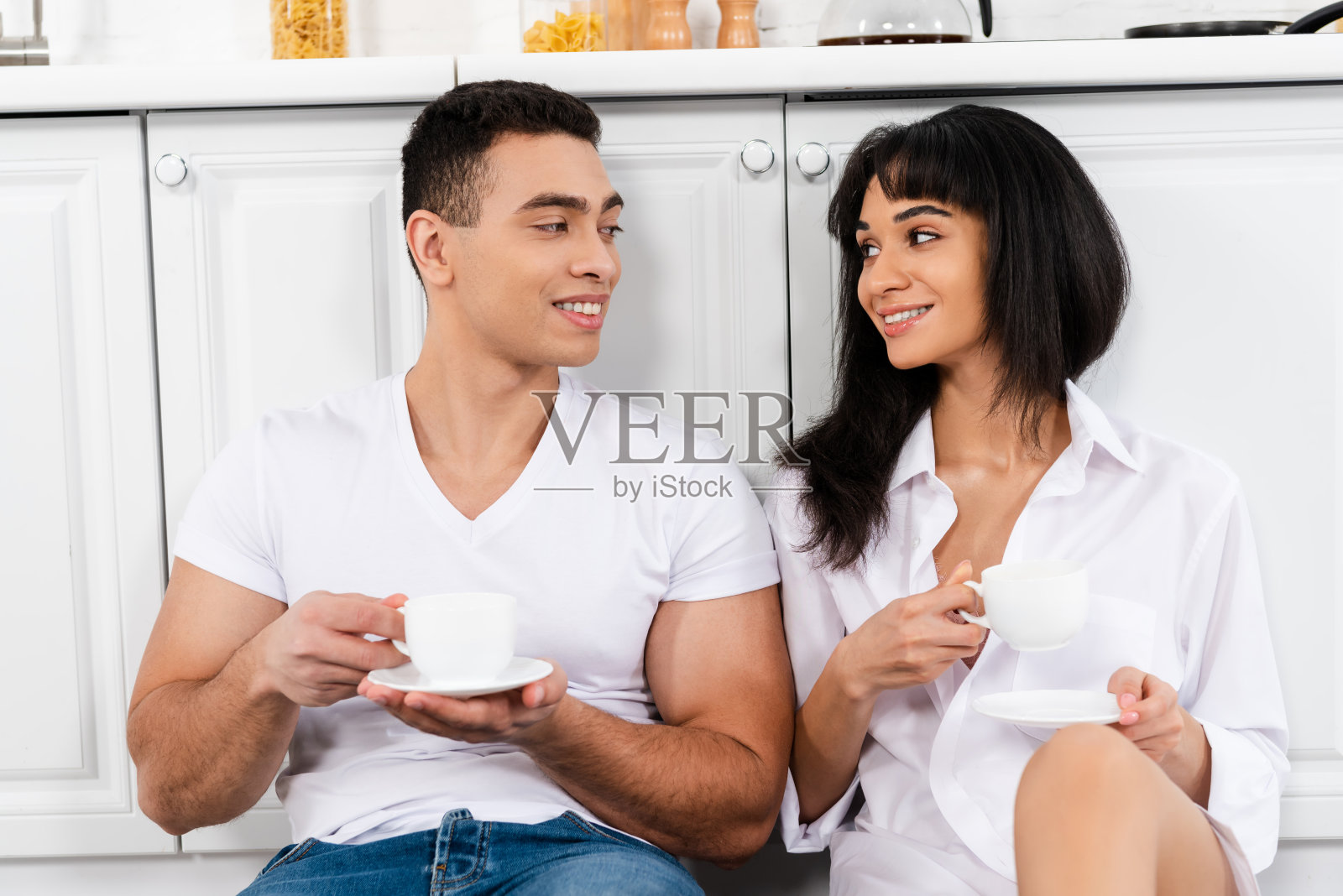 拥有茶托和咖啡杯的异族夫妇微笑着，在厨房橱柜旁边看着对方照片摄影图片