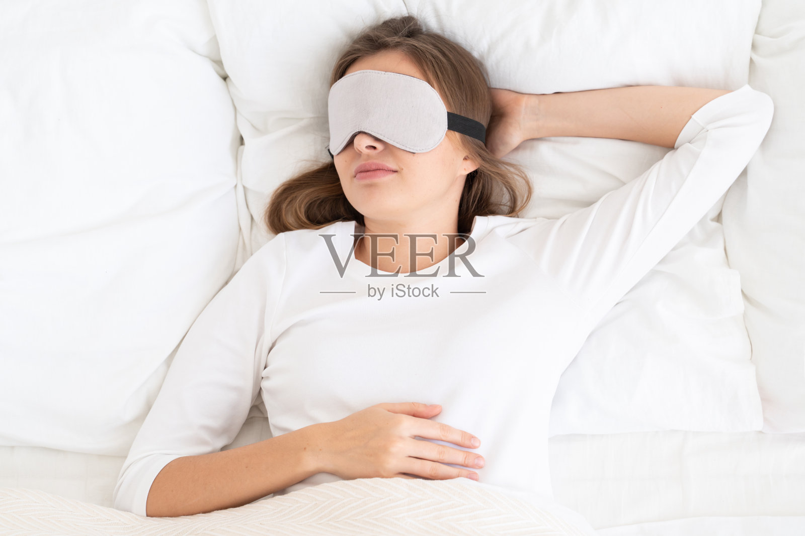 年轻的女孩躺在她的床上，白色的床单，戴着灰色的眼罩隔离她的阳光，安静地睡觉，放松照片摄影图片