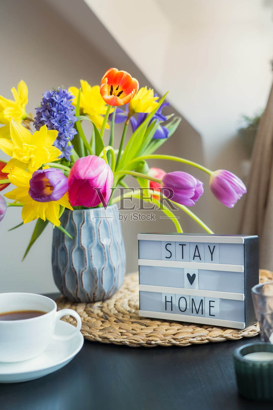 窗边的咖啡桌上放着一杯茶，一个写着“呆在家里”的灯箱和一束春天的鲜花。一切都是为了平静地呆在家里。有选择性的重点。垂直的名片。照片摄影图片