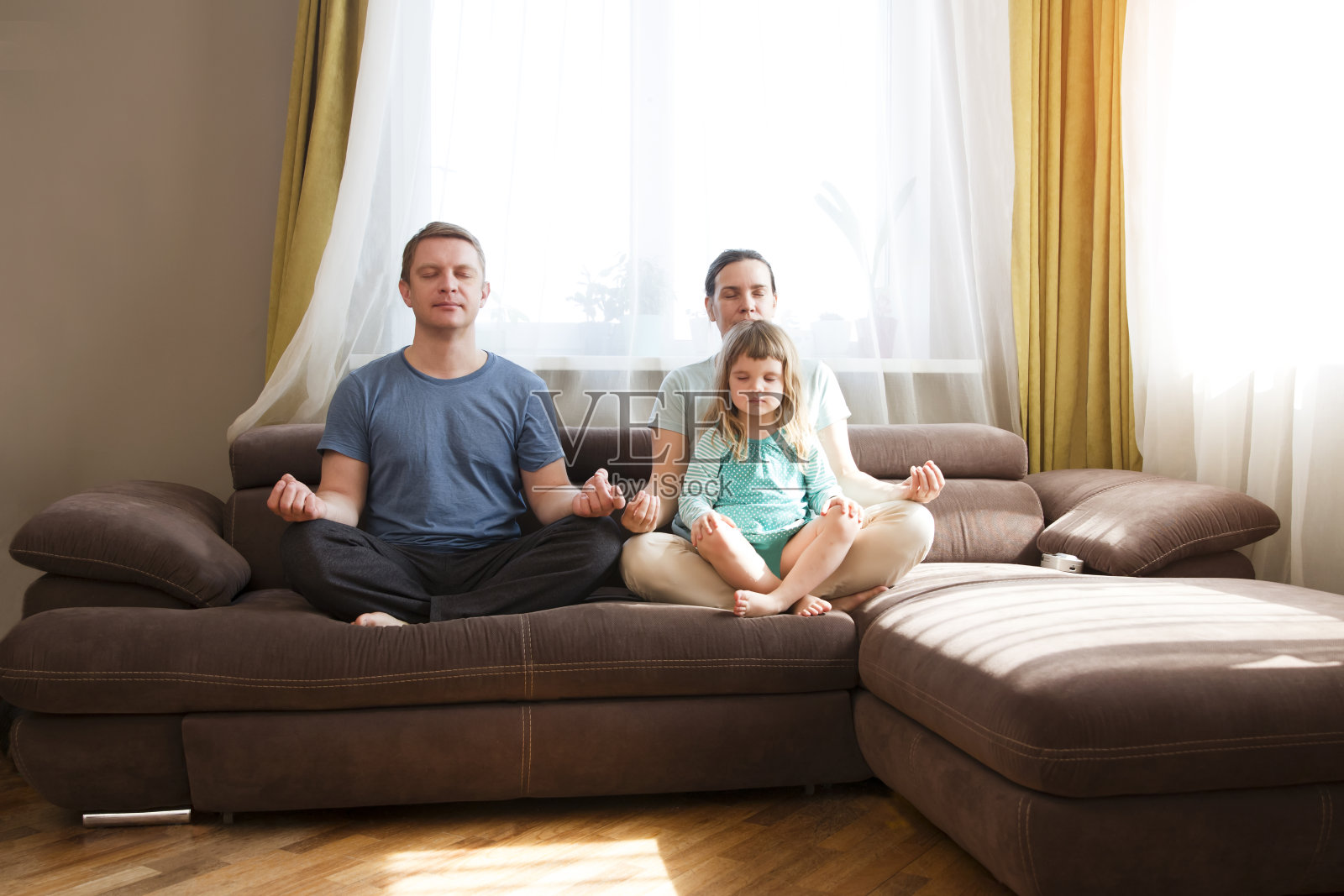 一家人和上幼儿园的女儿坐在沙发上一起练瑜伽。父母冥想缓解负面情绪周末在家隔离。照片摄影图片