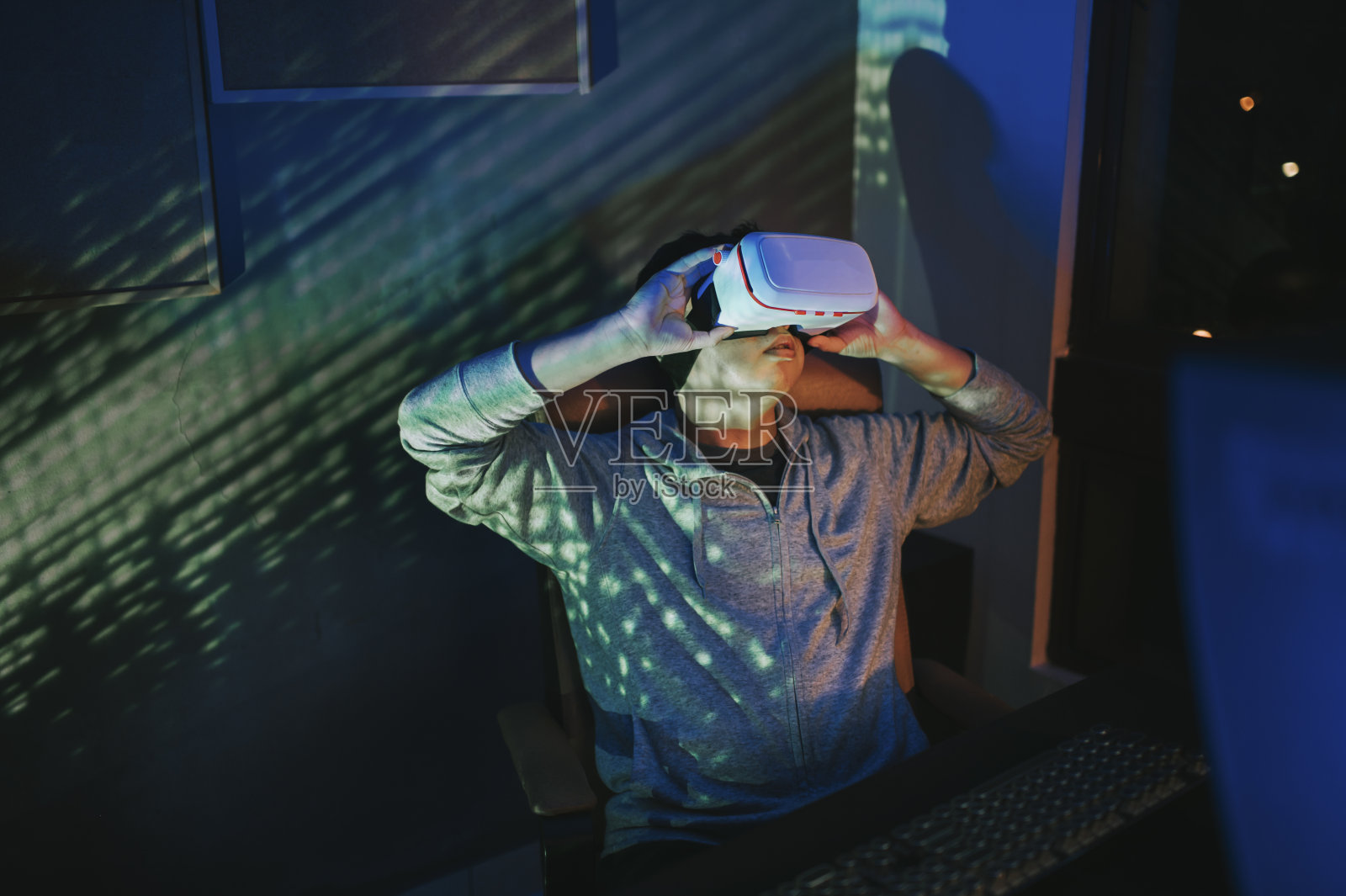 一名亚裔中国男性，晚上在他的家庭办公室自习室，戴上VR眼镜，在他的台式电脑前体验3D虚拟游戏照片摄影图片