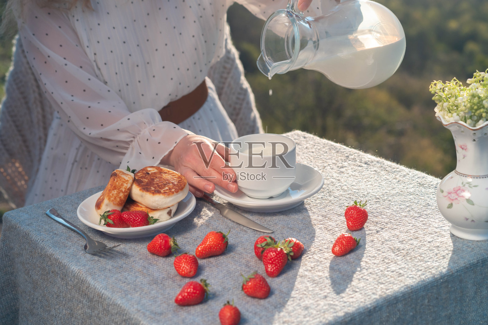 女人手里拿着一个盛满牛奶的杯子，放在放着奶酪蛋糕的桌子上。照片摄影图片
