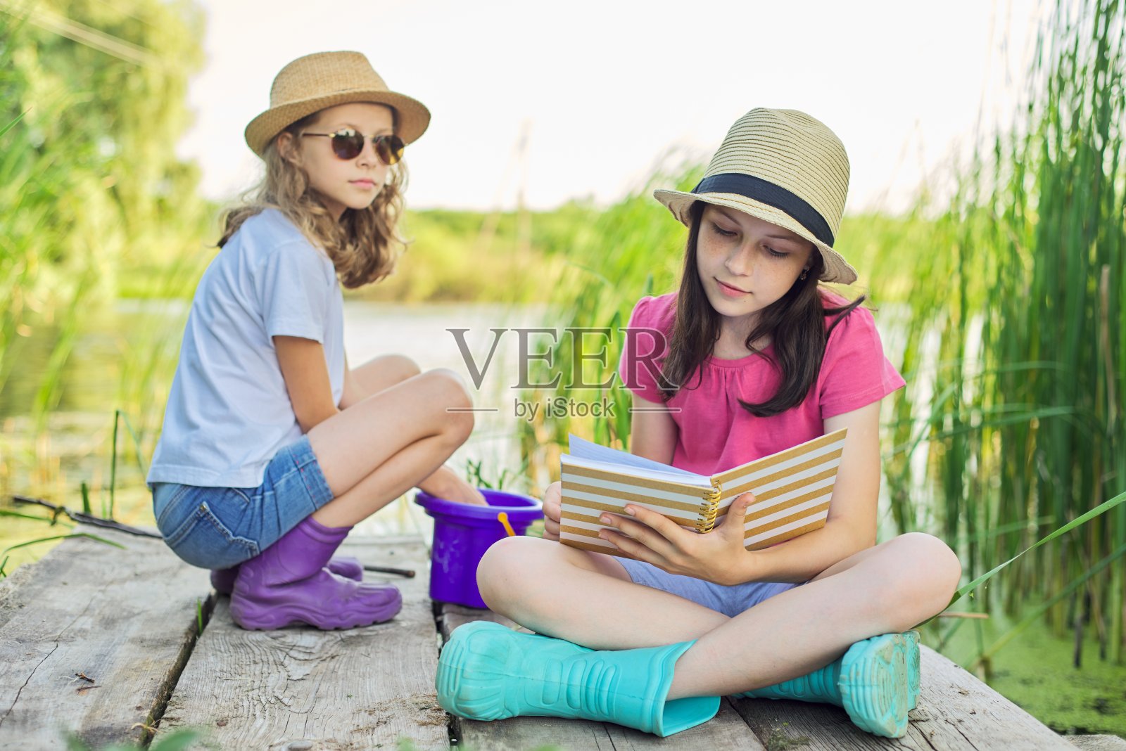 孩子们，两个女孩坐在湖边的木码头上，一边聊天一边看笔记本照片摄影图片