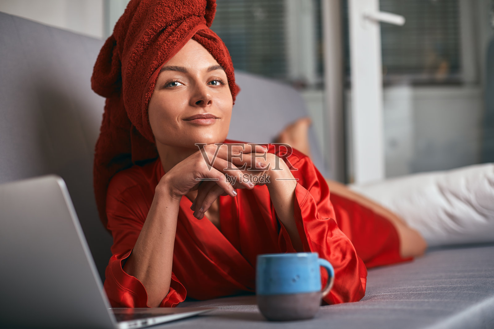 一名身穿红色浴衣、头戴红色毛巾的年轻女子正站在厨房靠近桌子的地方，喝着咖啡，使用着笔记本电脑。早上，女孩淋浴后喝茶和工作在电脑上。自由职业者在家工作照片摄影图片