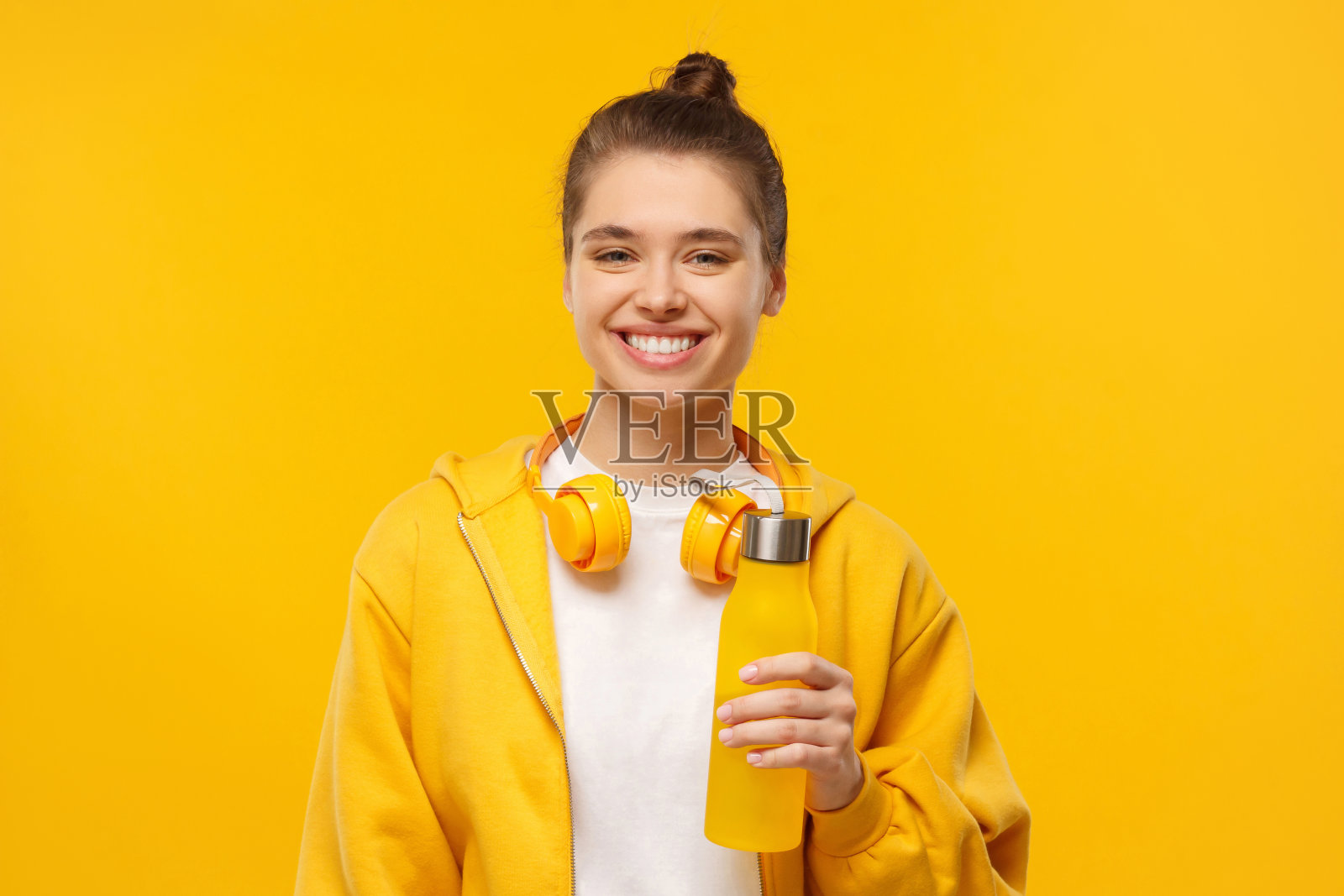 快乐女孩穿着t恤和帽衫，脖子上戴着无线耳机，手里拿着可重复使用的水瓶，孤立在黄色的背景上照片摄影图片
