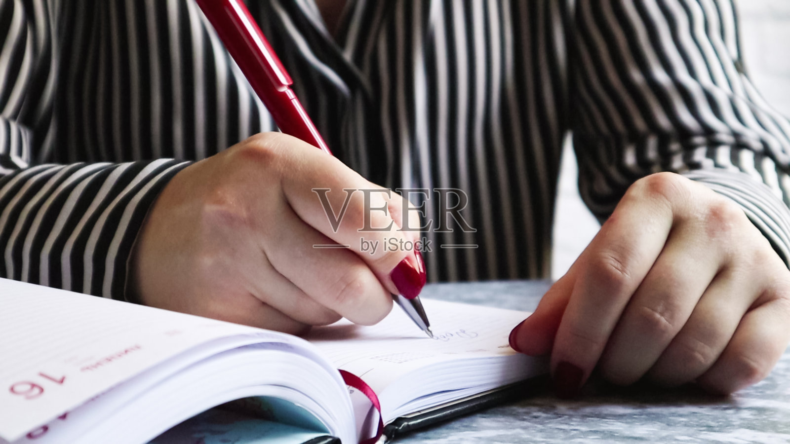 一个有着红色指甲的女人的手的侧面，拿着一支红色的笔，在一张空的日记本上写着什么，躺在一张灰色的桌子上。女孩在一个黑色的笔记本上做笔记照片摄影图片