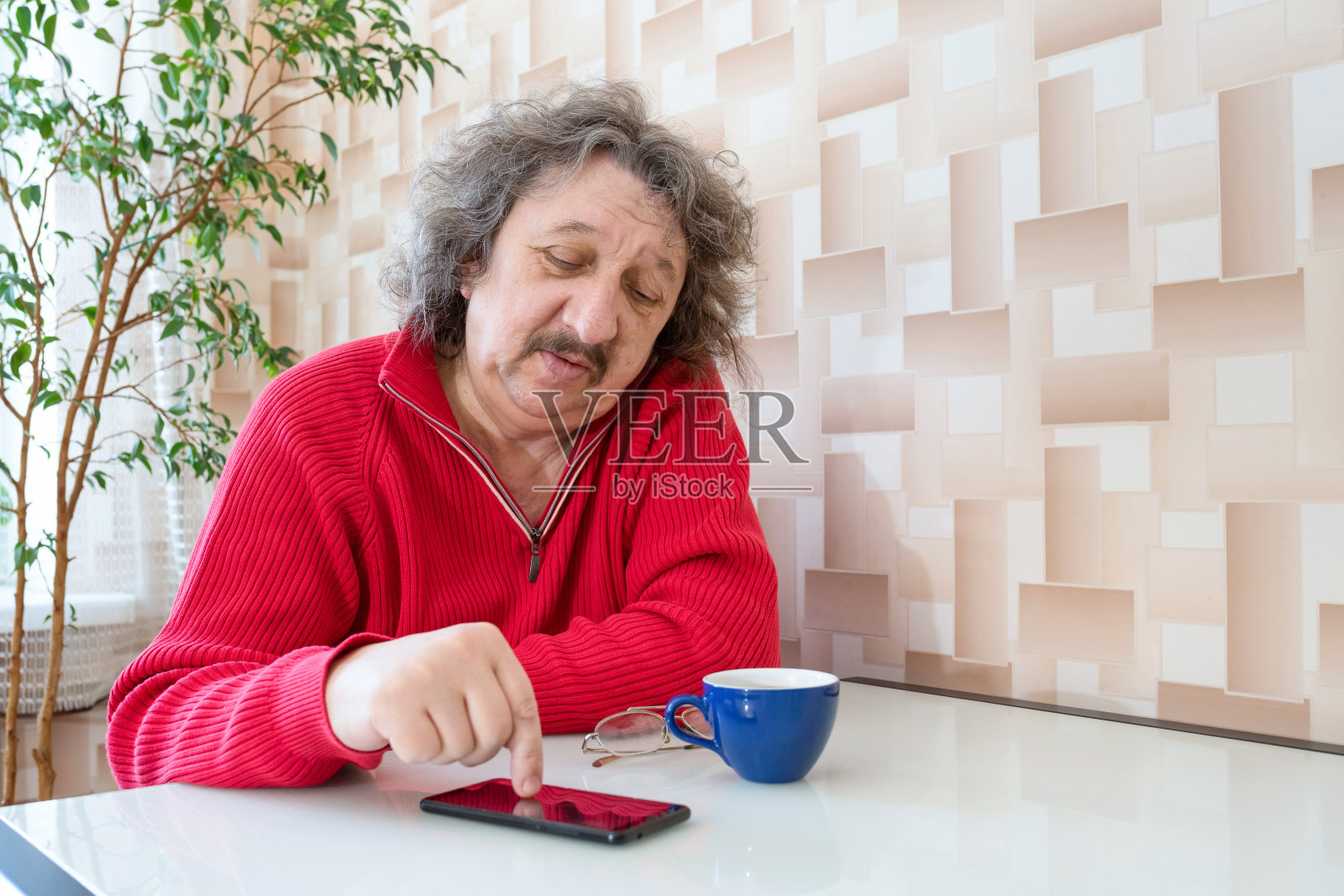 9. 一位身穿红色套头衫的老人，手里拿着电话坐在桌子旁。照片摄影图片