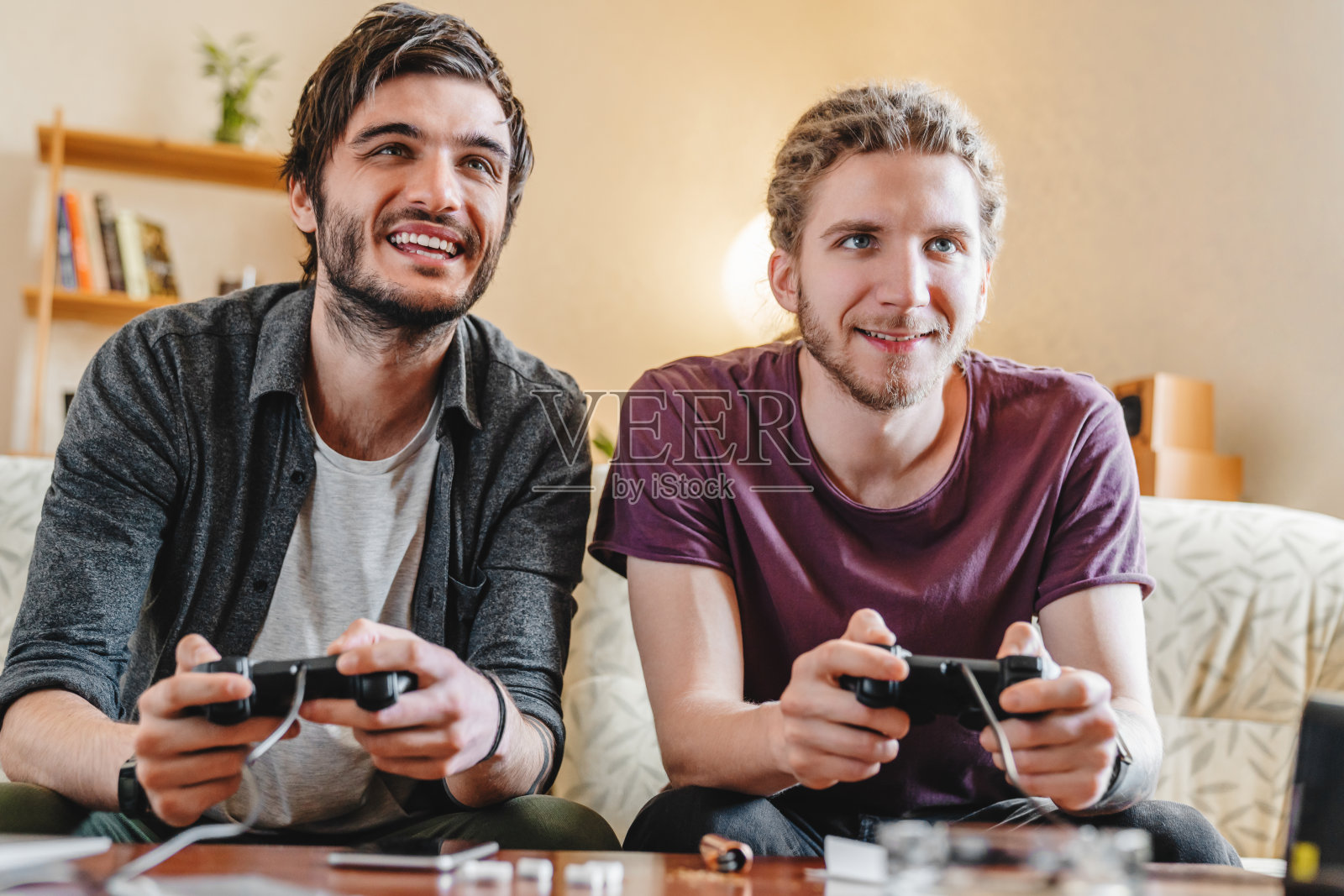 两个微笑的年轻人坐在家里的沙发上玩电子游戏照片摄影图片