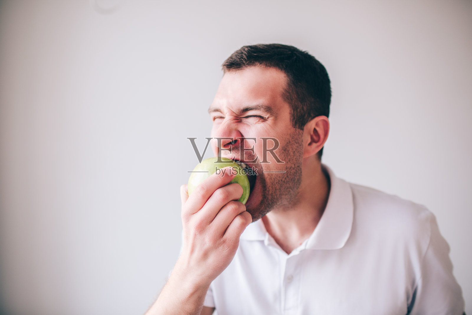 穿着白衬衫的年轻人在背景中显得孤立。小伙子咬着一块又绿又新鲜又好吃的苹果。好吃美味的水果。照片摄影图片