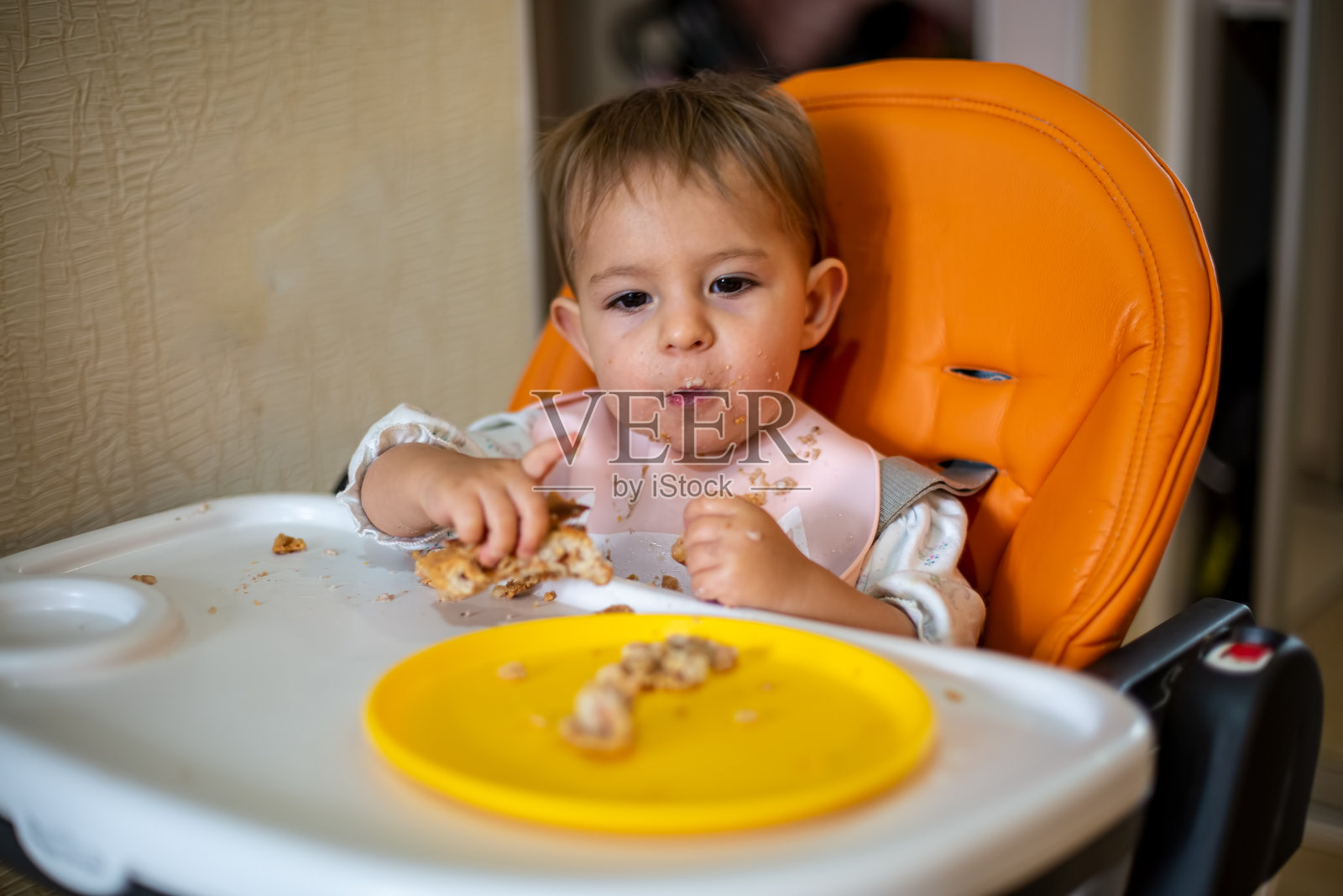 橙色婴儿座上的可爱宝宝选择一块蛋糕。面包屑，桌上放着一个橙子盘子和一个馅饼。特写，前视图，软焦点，模糊背景照片摄影图片