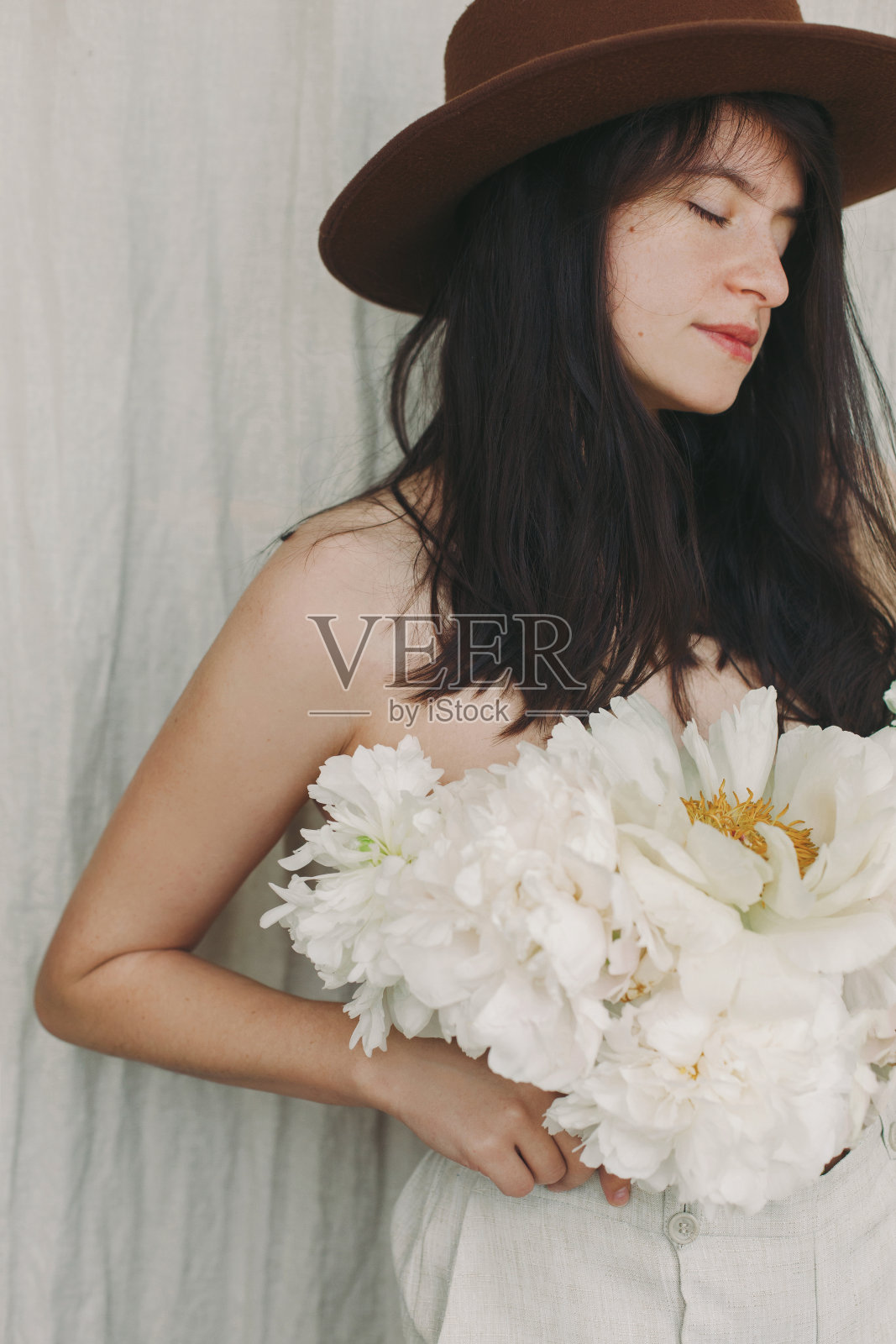 时髦的女孩戴着帽子与大的白色牡丹摆姿势。性感美女的形象与牡丹花束代替衬衫。清新芬芳的花香概念。自我护理和治疗照片摄影图片