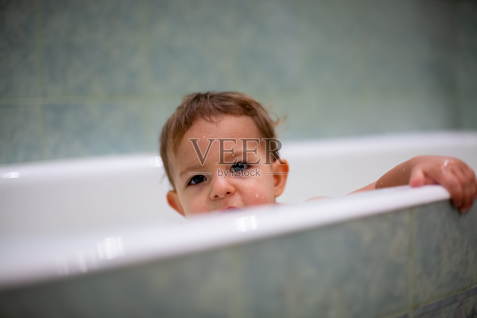 可爱顽皮，顽皮Ñ澳洲宝宝洗澡时，微笑着看镜头，把手放在浴缸的一边。背景是一间模糊的绿色浴室。特写,软焦点照片摄影图片
