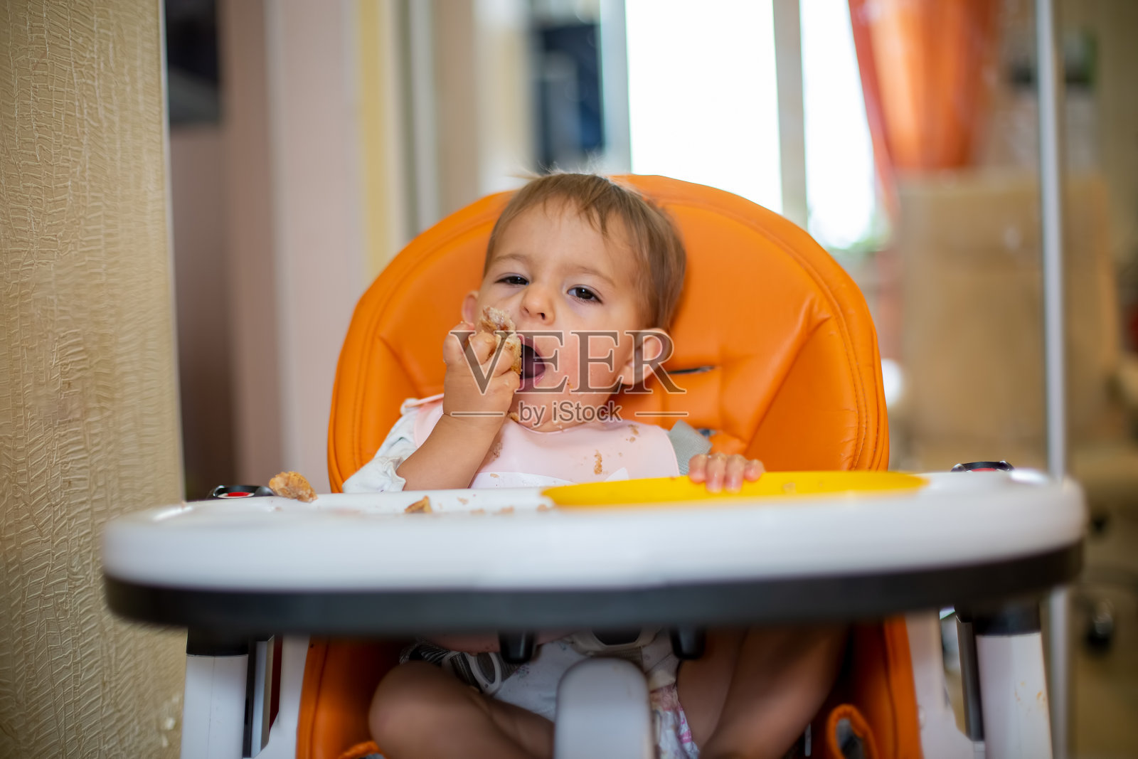 可爱的白人婴儿坐在一个橙色的婴儿椅上，用他的手从一个橙色的塑料盘子里吃一个派，并且看着镜头。特写，前视图，软焦点，模糊背景照片摄影图片