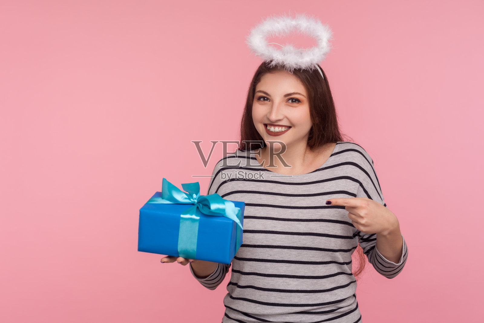 快乐天使女孩的肖像与圣人光环在头上指向生日礼物和微笑，持有礼物，捐赠照片摄影图片