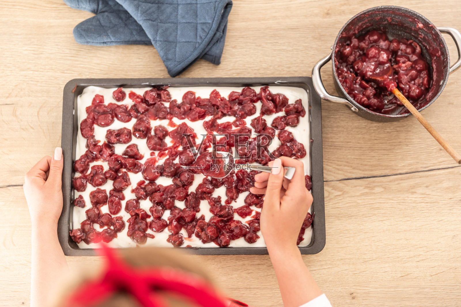 一个女人用勺子把煮熟的樱桃放在蛋糕的奶油糖霜上的俯视图。照片摄影图片