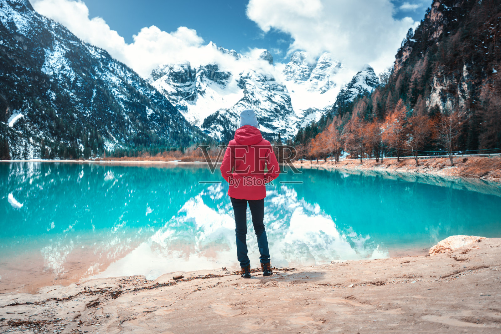 身着红夹克的年轻女子站在意大利多洛米特碧水的秋日湖畔。风景与女孩，倒影在水中，雪山，蓝天与白云，树木。旅行照片摄影图片