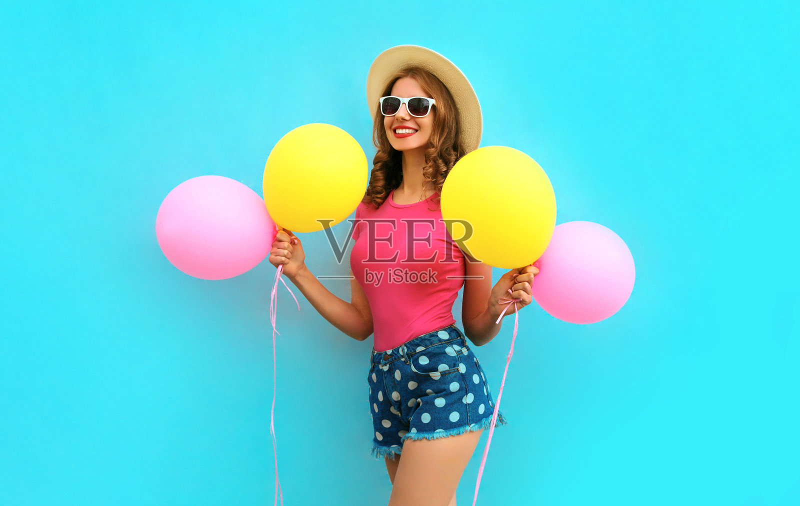 夏天彩色的愉快的图像快乐的年轻女子与黄色的粉红色气球有乐趣穿着短裤和草帽在蓝色的墙壁背景照片摄影图片