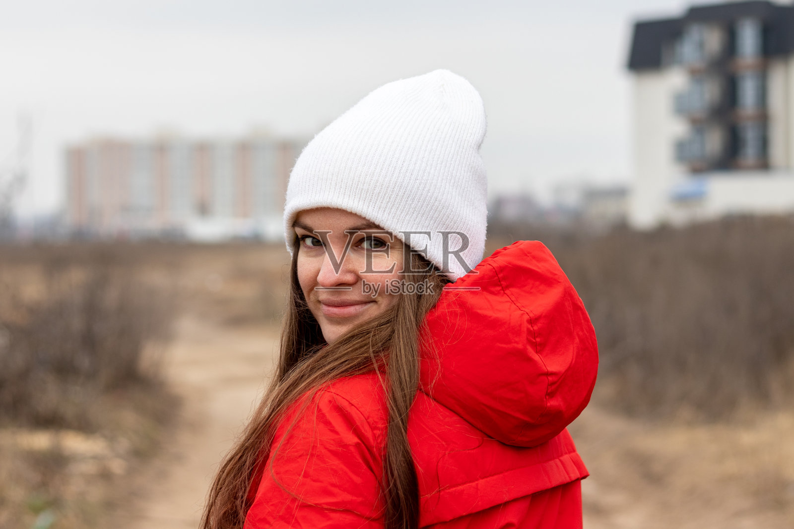 微笑的年轻女子与长头发在白色针织帽和红色夹克兜帽看在她的肩膀在阴天的街道上模糊的背景。女性素颜，自然美照片摄影图片