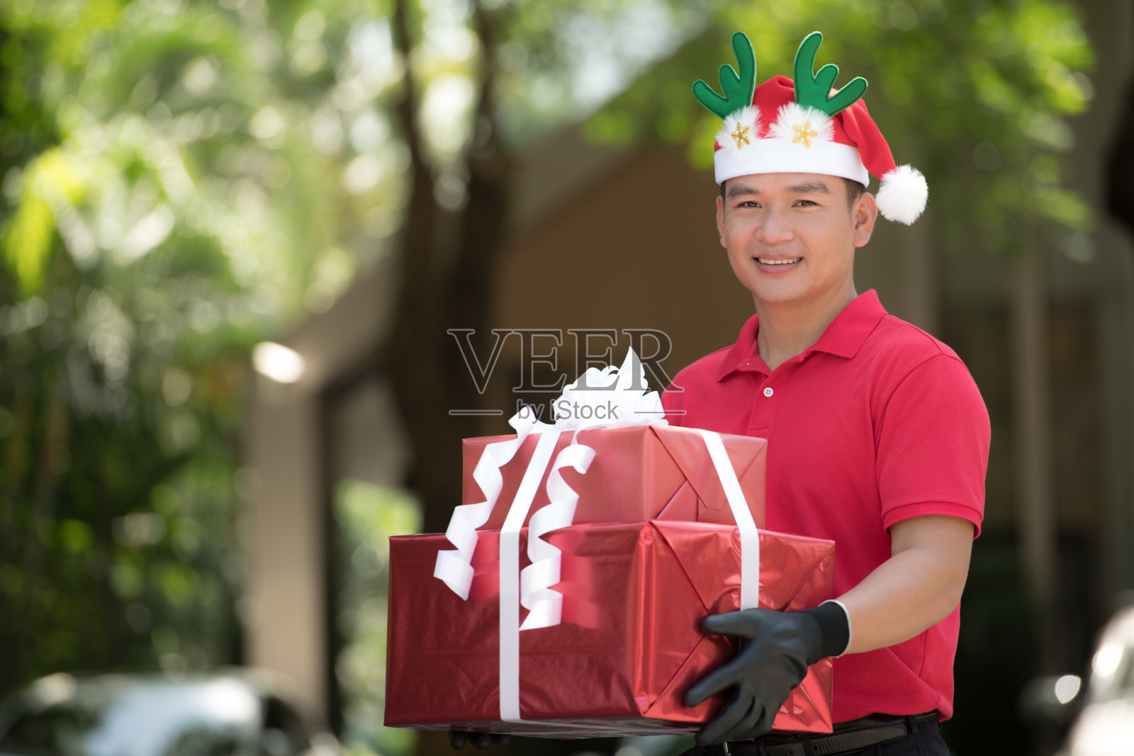一名身穿红色制服、头戴圣诞帽的亚洲快递员，在圣诞节期间为收件人递送礼物和礼品盒照片摄影图片