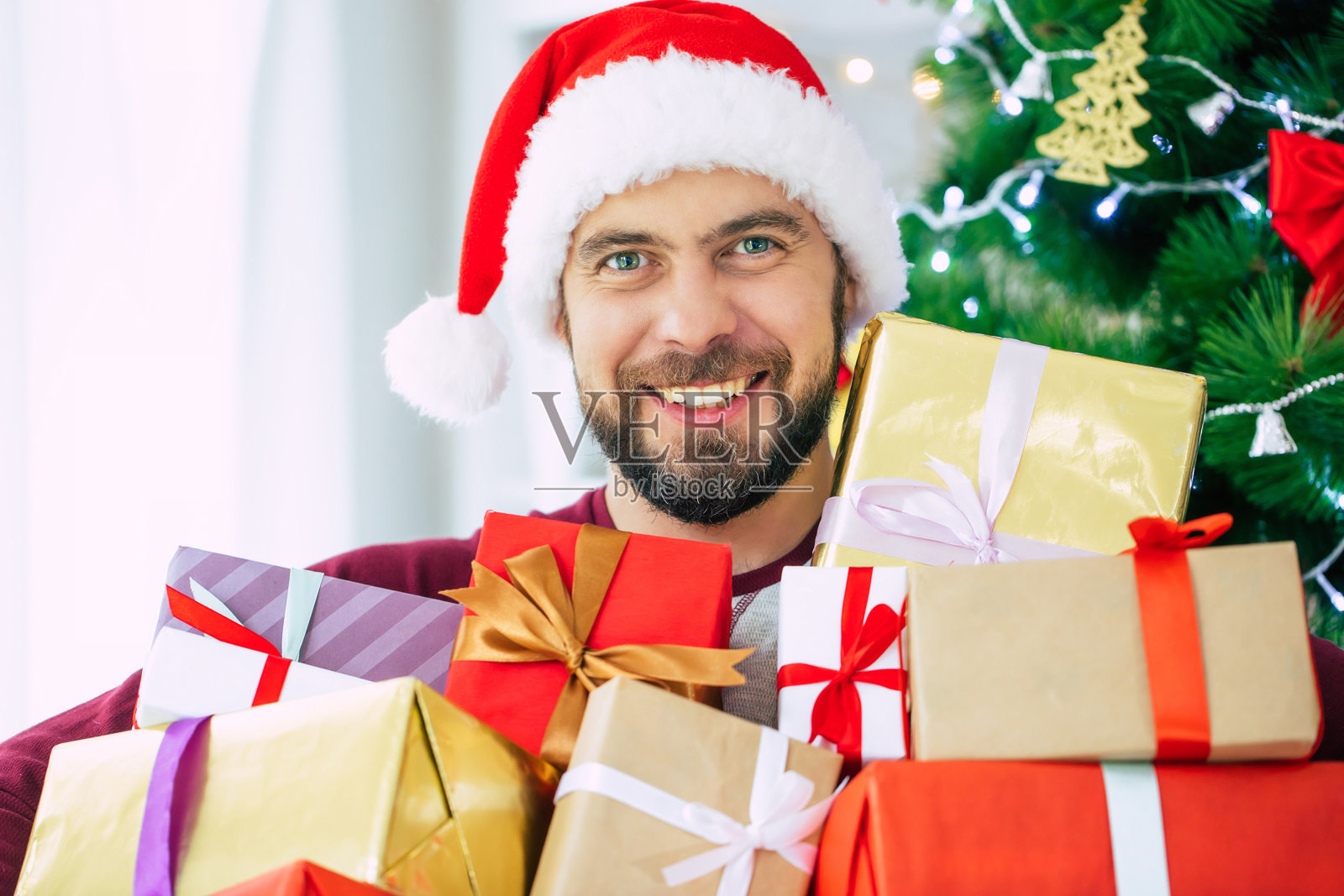 英俊兴奋的大胡子年轻人戴着圣诞帽，手里拿着许多礼物，在圣诞树的背景上摆姿势照片摄影图片