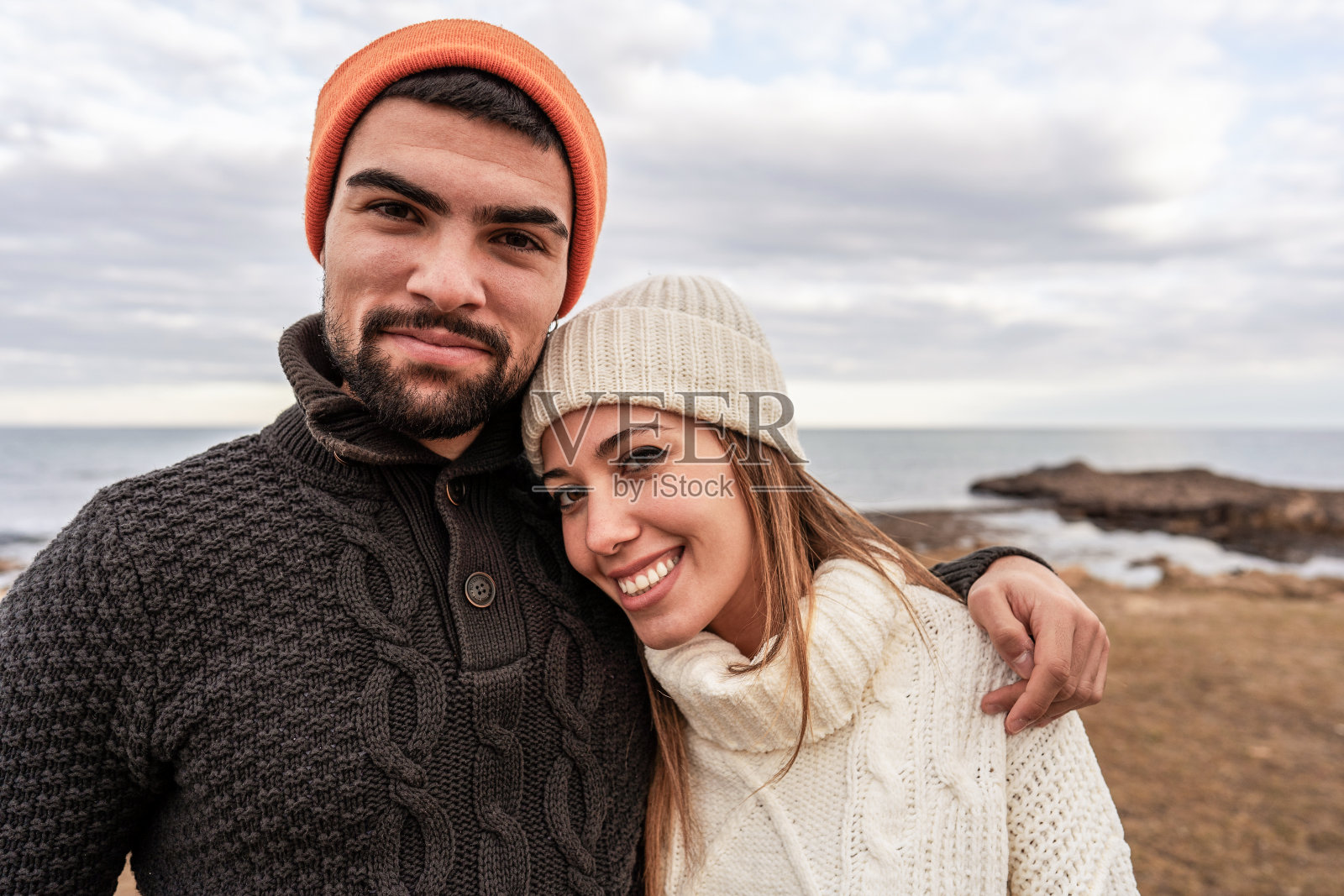 美丽的年轻夫妇相爱站在海边度假看相机摆姿势肖像与多云的天空在冬季海上度假-白种女友休息她的头在他的男朋友的肩膀上照片摄影图片