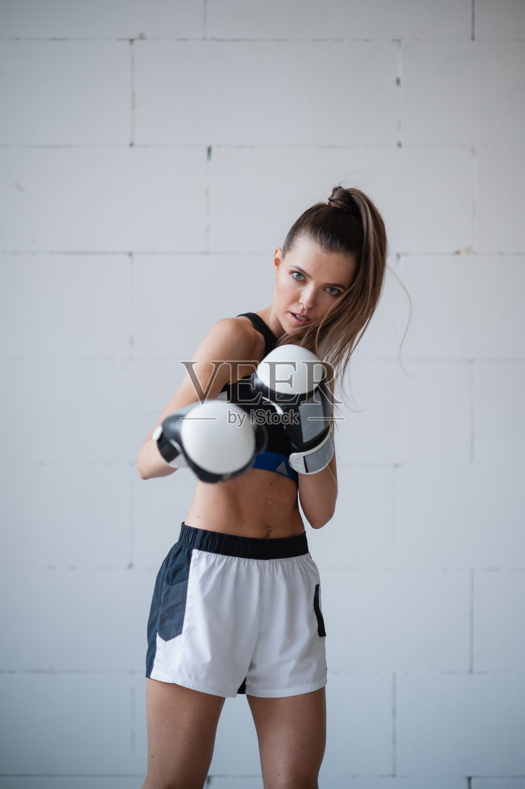 一位漂亮的女运动员戴着拳击手套，穿着运动服，靠着一堵白砖墙站着照片摄影图片
