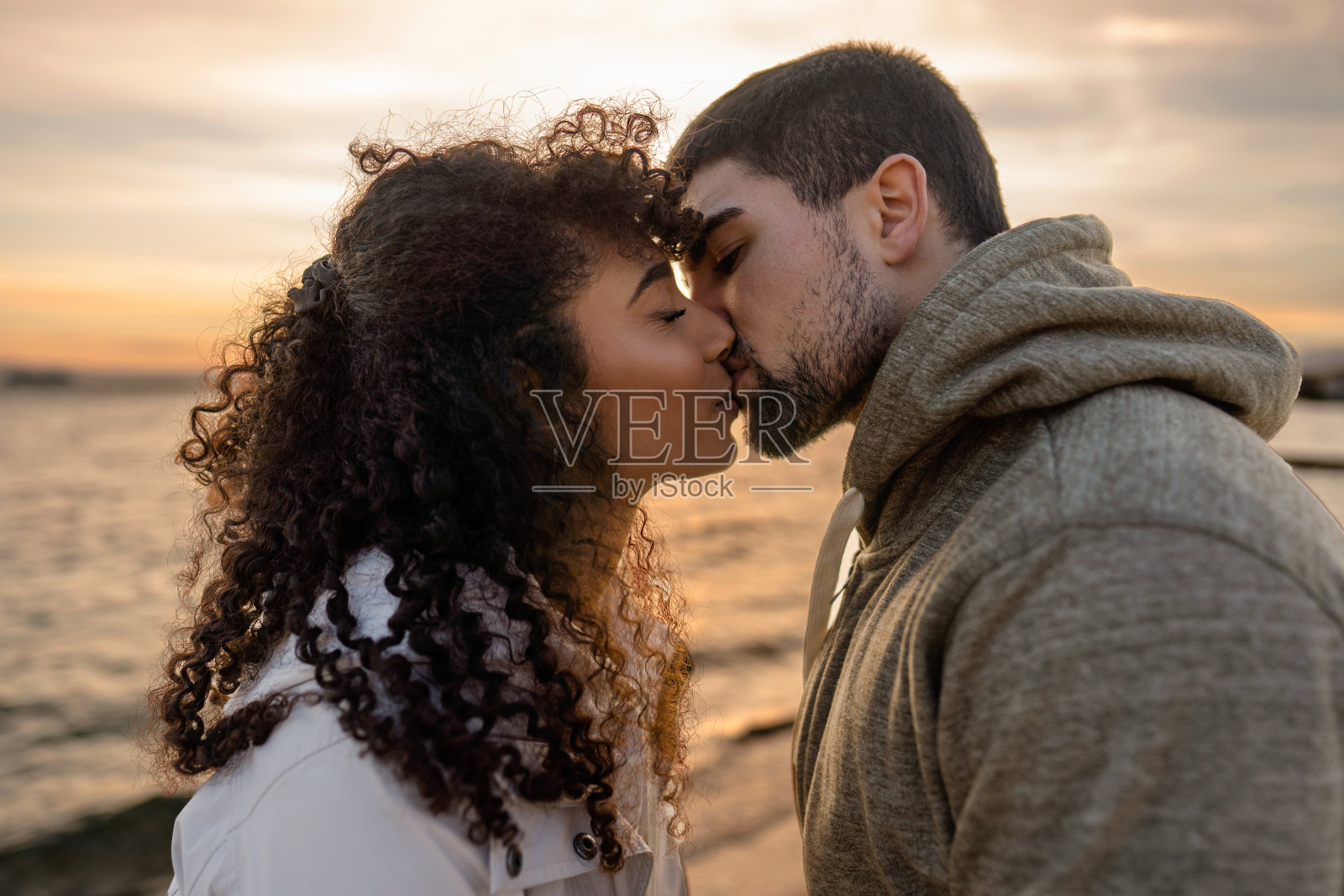 头和肩膀的肖像年轻美丽的夫妇在冬季海边度假胜地与多云的天空在日落亲吻。两个千禧一代在度假旅行中，在户外表达他们的心声照片摄影图片