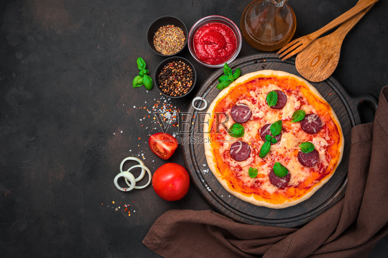 在混凝土背景下，棕色切菜板上放着意大利辣香肠披萨，配料和餐巾照片摄影图片