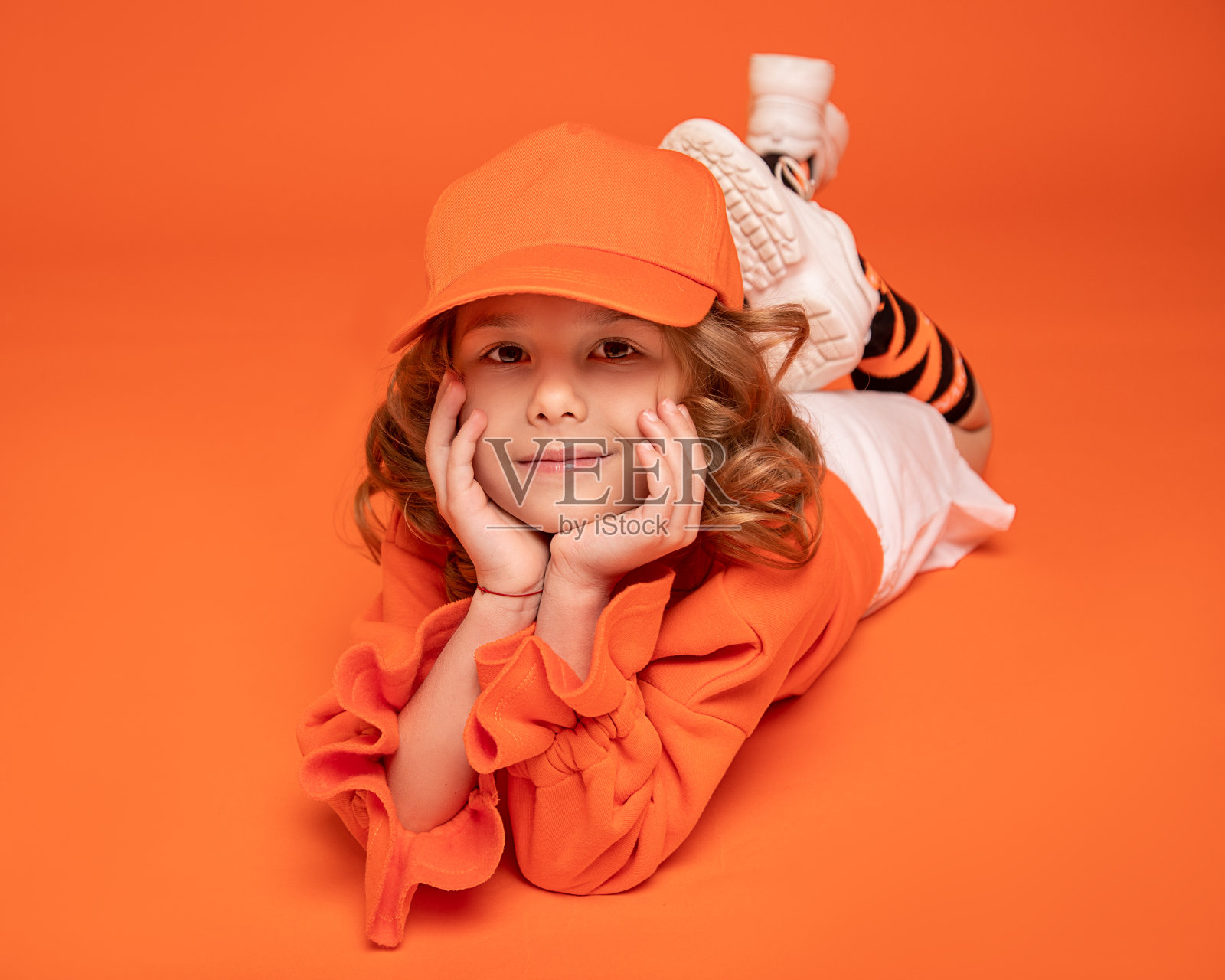 小女孩戴着帽子，穿着时髦的衣服，微笑着躺在工作室的地板上，橙色的背景。模拟了照片摄影图片