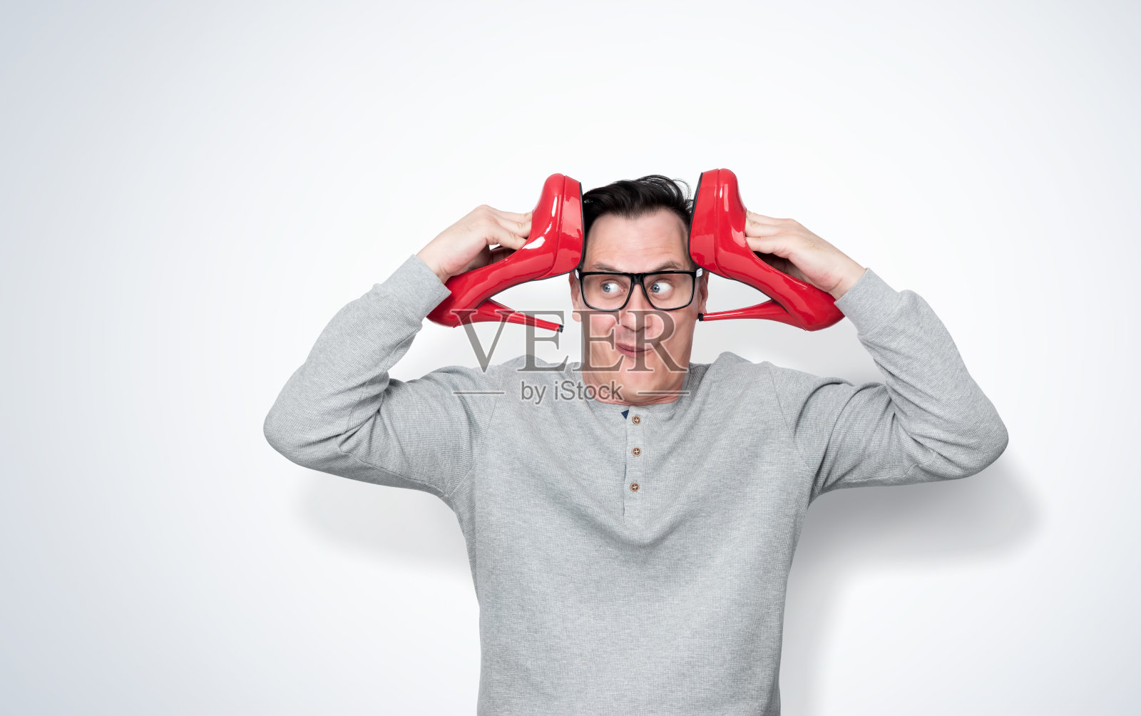 一个滑稽有趣的男人戴着眼镜，双手拿着女性红色高跟鞋靠近他的头，在灯光背景。照片摄影图片