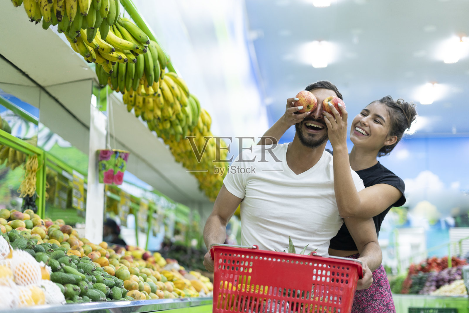 一对拉丁夫妇在一家杂货店里购物，妻子用两个苹果遮住她伴侣的眼睛照片摄影图片