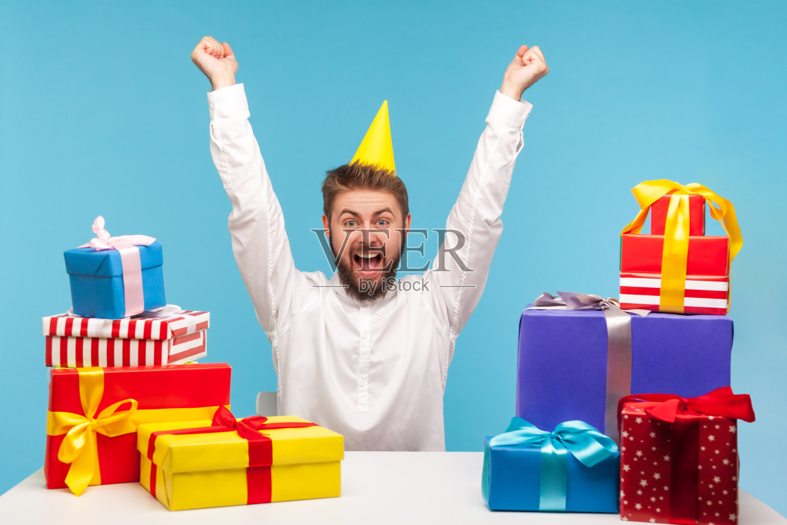 快乐快乐的男人留着大胡子戴着圆锥帽尖叫着举起双手，庆祝自己的生日，坐在家里的办公室里收到许多五颜六色的礼盒照片摄影图片