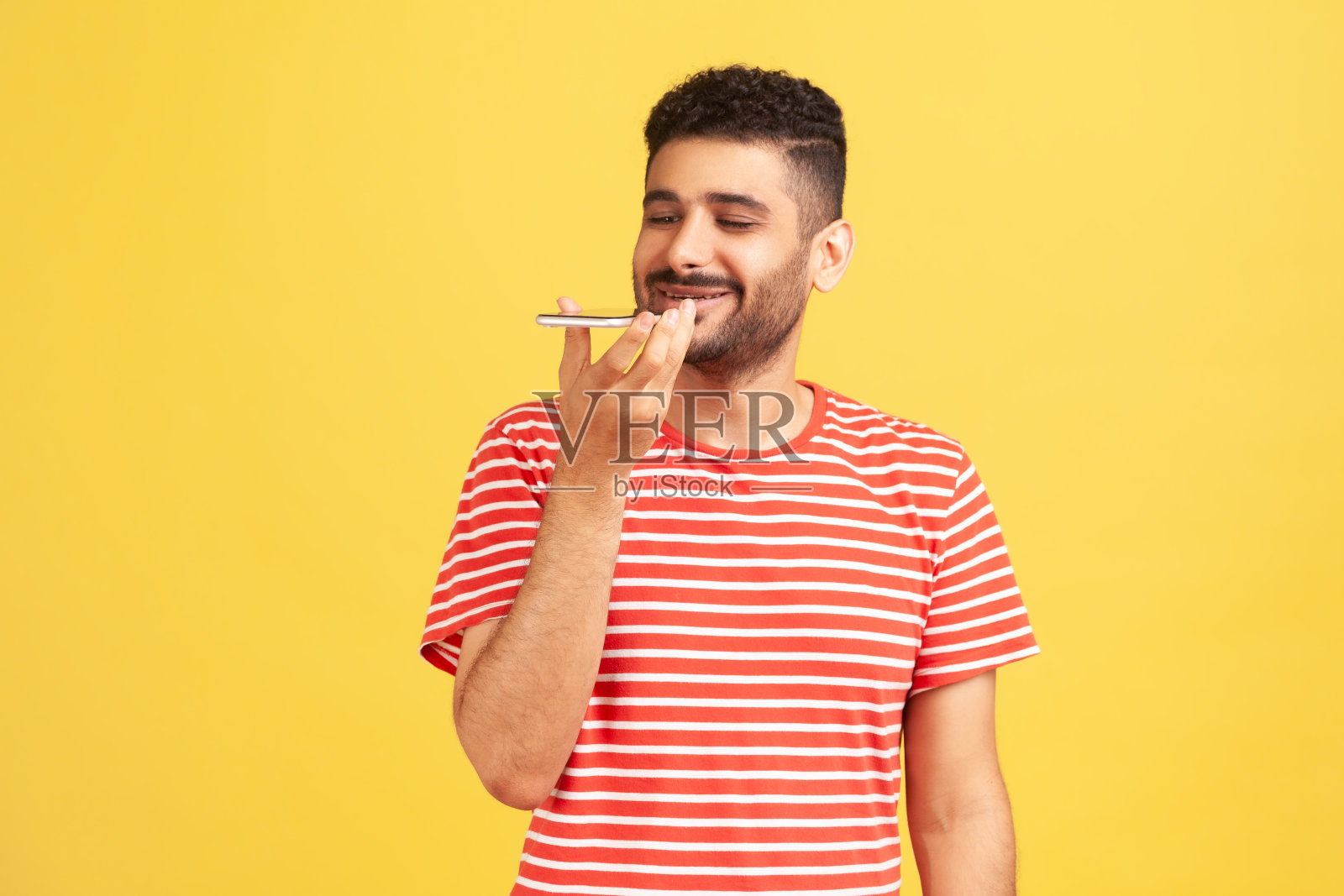 一个留着胡子、穿着条纹t恤的快乐满意男士，将手机拿在嘴边，用智能手机记录语音信息或与在线助手交谈照片摄影图片